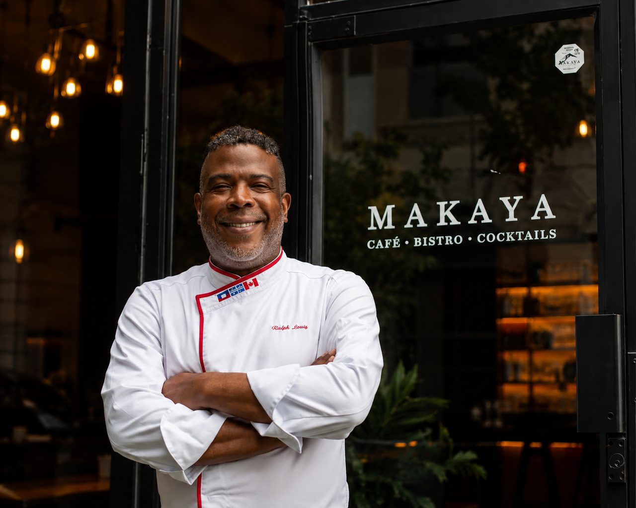 Ralph Leroy en veste blanche de chef, bras croisés et souriant, devant la porte vitrée du bistro Makaya au centre-ville de Montréal