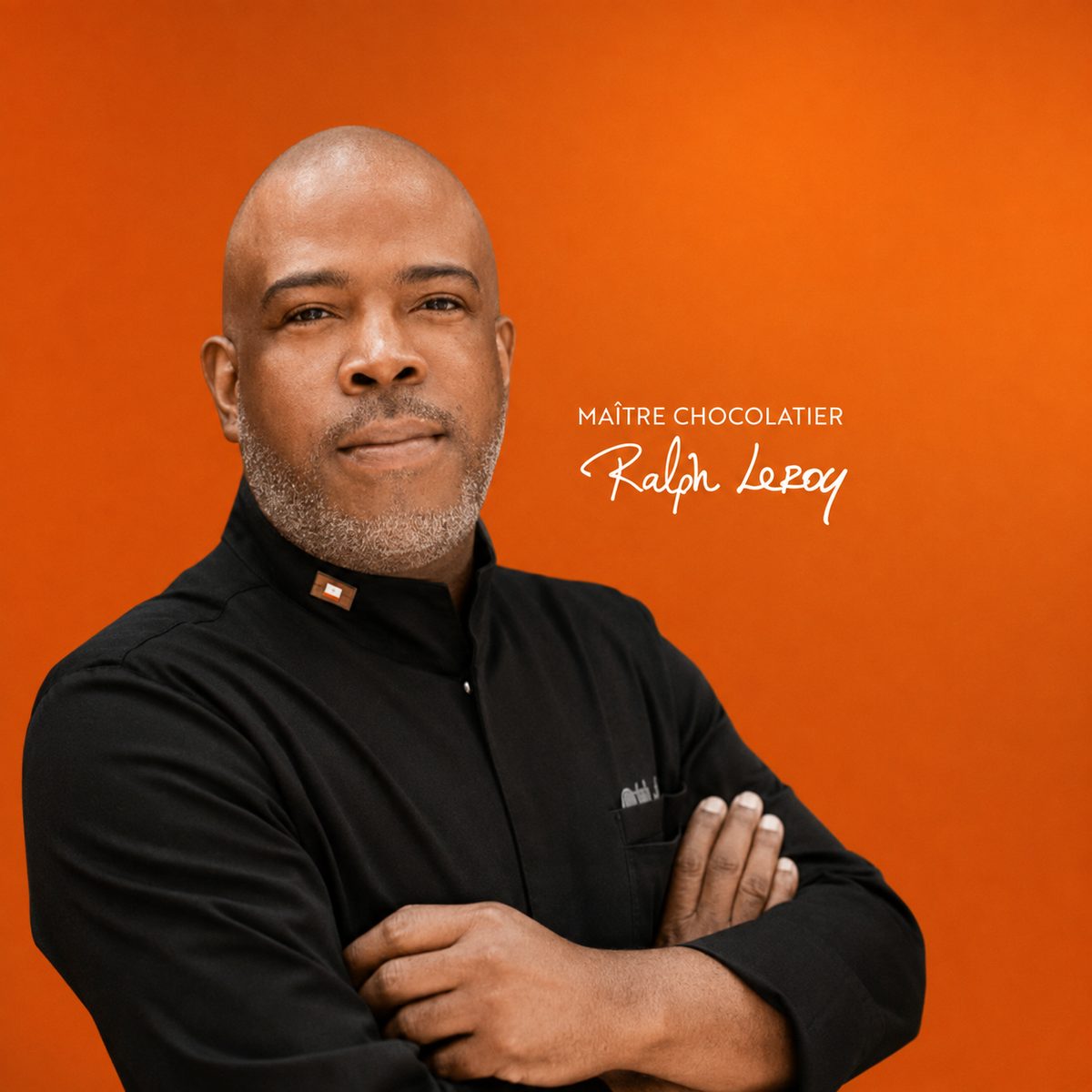 Portrait officiel de Ralph Leroy Prophète en veste noire de chocolatier, bras croisés, sur fond orange, avec sa signature