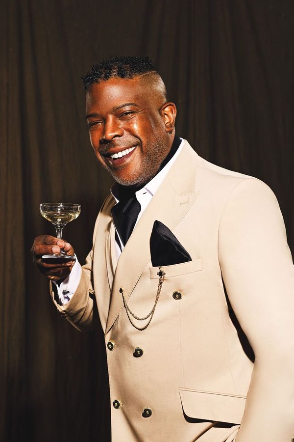 Ralph Leroy en costume croisé crème, pochette noire et coupe de champagne à la main, souriant sur fond de rideau sombre