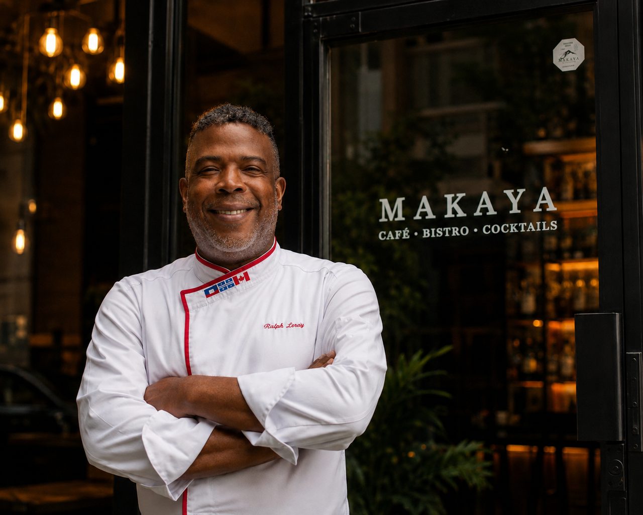Ralph Leroy souriant, bras croisés, en veste blanche de chef brodée à son nom et aux drapeaux du Québec et du Canada, devant la porte du bistro Makaya